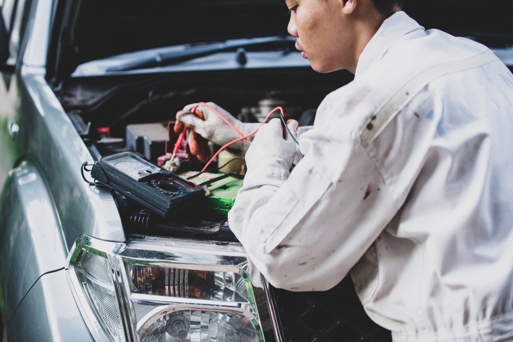 A mechanic in a white uniform uses a multimeter to test the battery of a car with an open hood, showcasing the importance of thorough car services in Singapore.