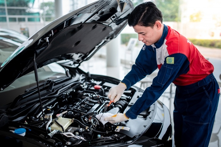 A mechanic wearing a blue and red uniform works on an engine of a car with an open hood in a service center, expertly performing car repairs including brake pad replacement.