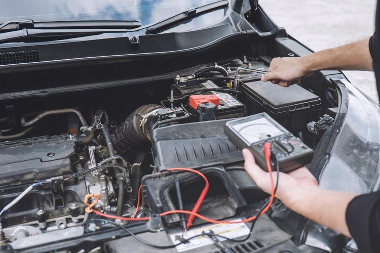 A person uses a multimeter to check the battery of a car, with the car's hood open and engine visible, ensuring there are no transmission problems.