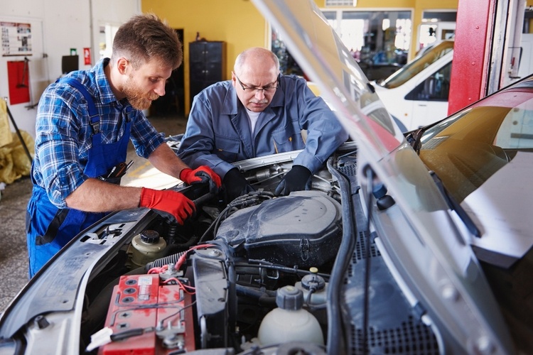 Two mechanics work together under the hood of a car in a garage, addressing transmission problems. One wears a blue shirt and red gloves, while the other sports a grey uniform and black gloves.