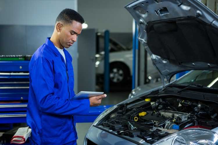 A mechanic in a blue uniform is inspecting a car engine with the hood raised, using a digital tablet for reference, likely checking for transmission problems as part of routine car maintenance in Singapore.